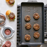 Burger patties on a grill pan with burgers and ingredients on a countertop.