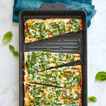 Rectangular pizza with spinach and cheese on black grill pan, surrounded by basil leaves.