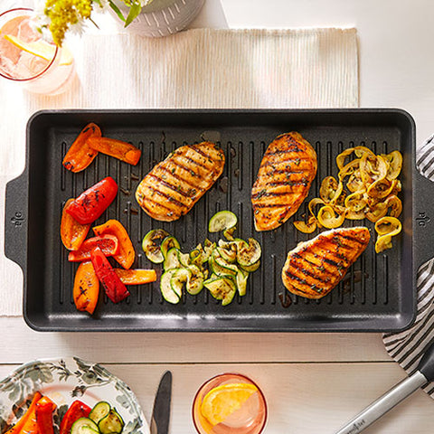 Grilled chicken and vegetables on a black, double burner grill pan with drinks and cutlery in the background.
