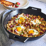 Large, black cast iron skillet with hash browns, eggs, and ham on a stone surface with toast and strawberries in the background.