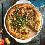 Pizza with arugula and other toppings in large, white, round pizza stoneware dish on a wooden table.