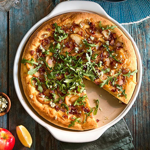 Pizza with arugula and other toppings in large, white, round pizza stoneware dish on a wooden table.