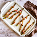 Bacon-wrapped asparagus on a white ceramic stoneware serving and cooking tray with a brown napkin.