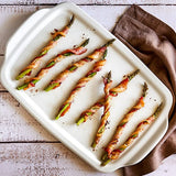 Bacon-wrapped asparagus on a white ceramic stoneware serving and cooking tray with a brown napkin.