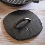 Black, cast iron grill press on a wooden surface with a grill pan in the background.
