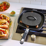 Black cast iron skillet, with grill press, on a stove with sausages cooking, next to a plate of hot dogs and vegetables.