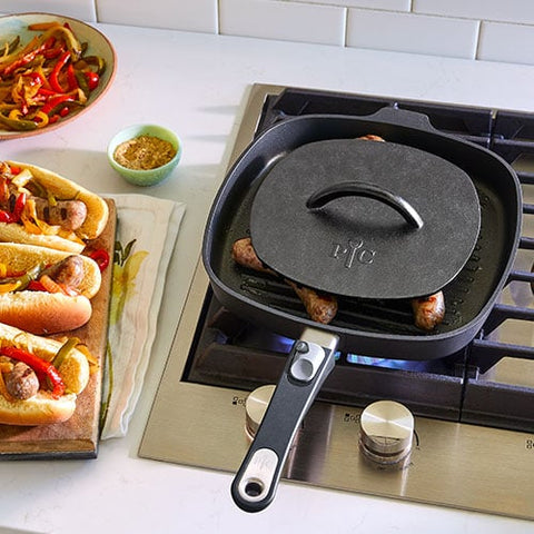 Black cast iron skillet, with grill press, on a stove with sausages cooking, next to a plate of hot dogs and vegetables.