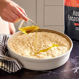 Person pouring olive oil onto dough in round baking pan with a Focaccia package in the background.
