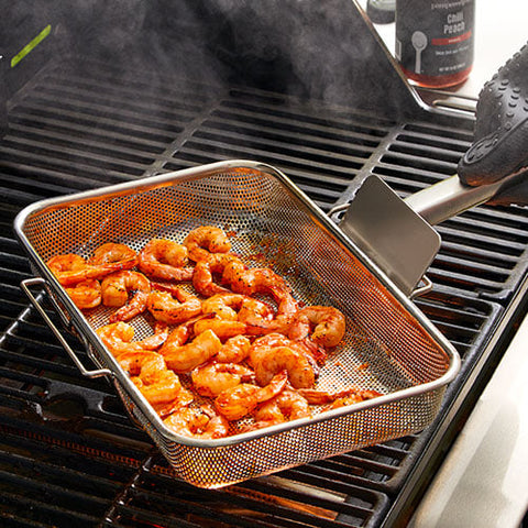 Shrimp being cooked in a mesh pan on a grill with a bottle of sauce in the background.