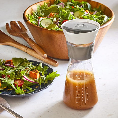 Vinaigrette salad dressing in a bottle with a wooden salad bowl and utensils on a white surface.