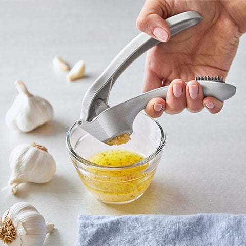 Person using garlic press to crush garlic into a glass bowl with garlic cloves on a light surface.