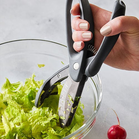 Hand using kitchen shears to cut lettuce in a glass bowl on a light surface.