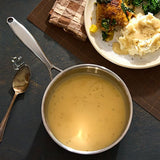 Metal pot with cheese sauce on a dark surface with a plate of food and checkered cloth.