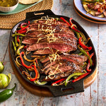 Steak with peppers and onions in a cast iron skillet on a wooden trivet board.