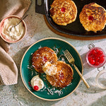Pancakes with whipped cream and cherries on a green plate with a Cast Iron Skillet and wooden trivet to the side.