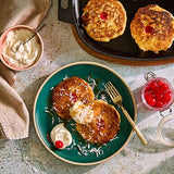 Pancakes with whipped cream and cherries on a green plate with a Cast Iron Skillet and wooden trivet to the side.