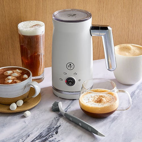 White, electric milk frother with various beverages on a marble surface.
