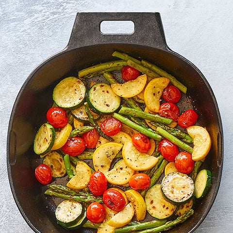 Vegetables including asparagus, zucchini, and tomatoes in a black, cast iron skillet on a light gray background.