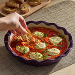 Person dipping bread into Italian Plum Stoneware Pie Plate of tomato-based dip with herbs on a textured surface.