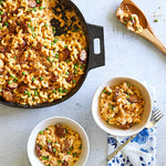 Macaroni and cheese with peas in a black cast iron skillet, along with bowls on a light background.