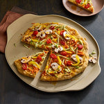 Vegetable pizza on a beige, large round pizza stoneware with a slice removed, on a dark surface.