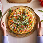 Person holding a pizza with vegetables on a large, round pizza stoneware.