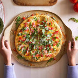 Person holding a pizza with vegetables on a large, round pizza stoneware.