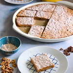 Sliced cake with powdered sugar on a white Large Round Pizza Stoneware Dish, surrounded by nuts and chocolate pieces on a light blue surface.