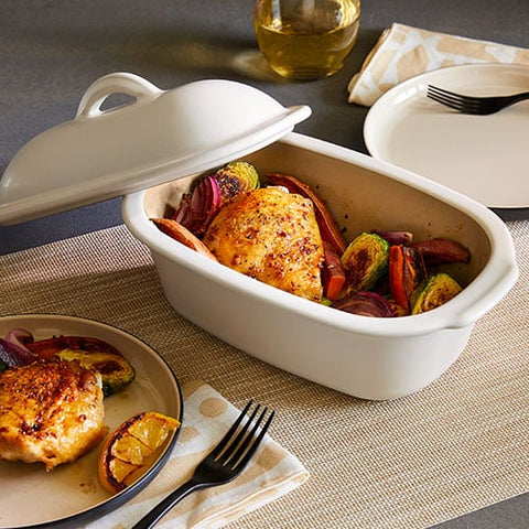 White, mini-deep stoneware covered baking dish with roasted chicken and vegetables on a table with a glass of wine.