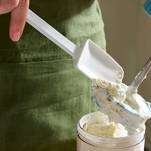 Person using a white mix and scraper spatula to scoop whipped cream into a container, wearing a green apron.