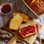 Sliced bread with red jam on a wooden cutting board, with a loaf of bread and a bowl of jam in the background.