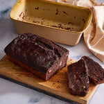 Loaf of chocolate bread with a slice cut, on wooden board with a Stoneware Bread Baking Loaf Pan and towel in the background.