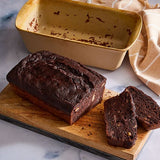 Loaf of chocolate bread with a slice cut, on wooden board with a Stoneware Bread Baking Loaf Pan and towel in the background.