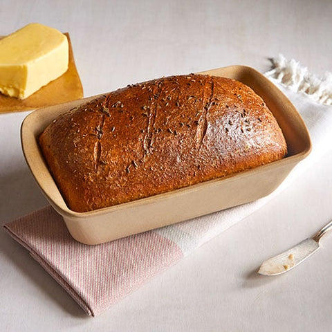 Loaf of bread in a beige, rectangular, natural stoneware bread baking loaf pan   on a light surface with butter and a knife.