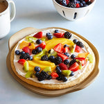 Fruit pizza on a Personal Pizza Stoneware with a bowl of berries and a cup of coffee in the background.