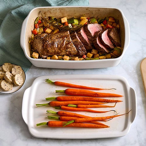 Roast beef with vegetables in a rectangular stoneware baking dish, accompanied by carrots on a tray cover.