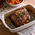 Roasted meat with herbs in rectangular stoneware baking and serving dish on a patterned tablecloth.