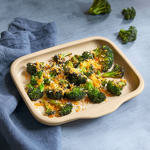 Beige stoneware bar pan with roasted broccoli and cheese on a blue surface with blue towel.