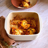 Baked dish with cheese and herbs in a square stoneware baking dish on a marble surface.