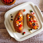 Two baked sweet potatoes with toppings on a white, square stoneware serving tray placed on a wooden surface.