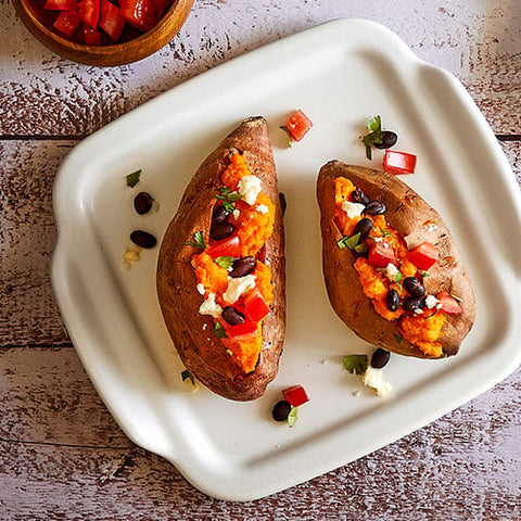 Two baked sweet potatoes with toppings on a white, square stoneware serving tray placed on a wooden surface.