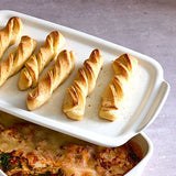 Baked breadsticks on a white rectangular stoneware serving and cooking tray with a dish of food underneath it. 