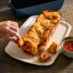 Bread loaf on stoneware serving tray with a hand taking a piece, next to a bowl of red sauce on a wooden surface.