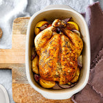 Roasted chicken with vegetables in White Satin Glazed Stoneware Covered Baking Dish on a wooden cutting board.