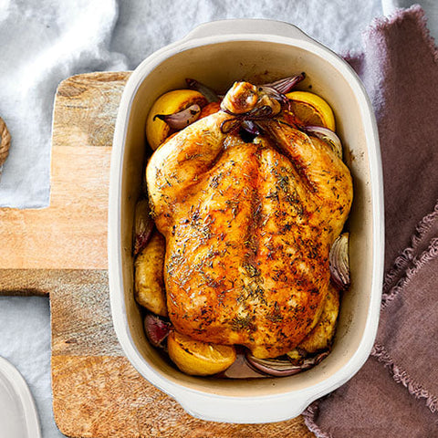 Roasted chicken with vegetables in White Satin Glazed Stoneware Covered Baking Dish on a wooden cutting board.