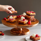 Wooden cake stand with dessert squares topped with whipped cream and raspberries, hand reaching for a piece.