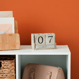 Wooden calendar on a white shelf against an orange wall.