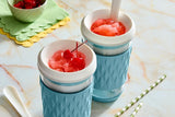 Two light blue, frozen drink maker tumbler cups filled with red slushie and a cherry, on a marble surface.