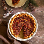 Pecan pie on a stoneware pie baking dish with decorative pine cones and a brown cloth on a wooden surface.