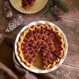 Pecan pie on a stoneware pie baking dish with decorative pine cones and a brown cloth on a wooden surface.